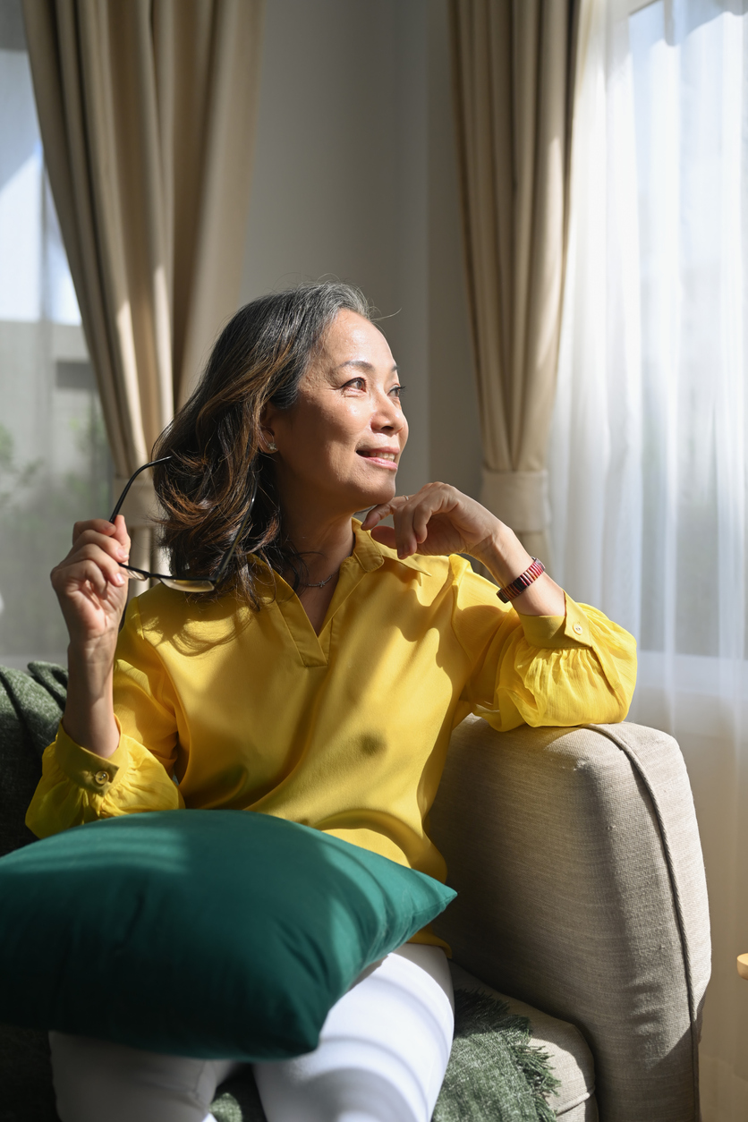 Happy middle aged woman resting on sofa and looking through the window, enjoy stress free peaceful mood wellbeing at home.